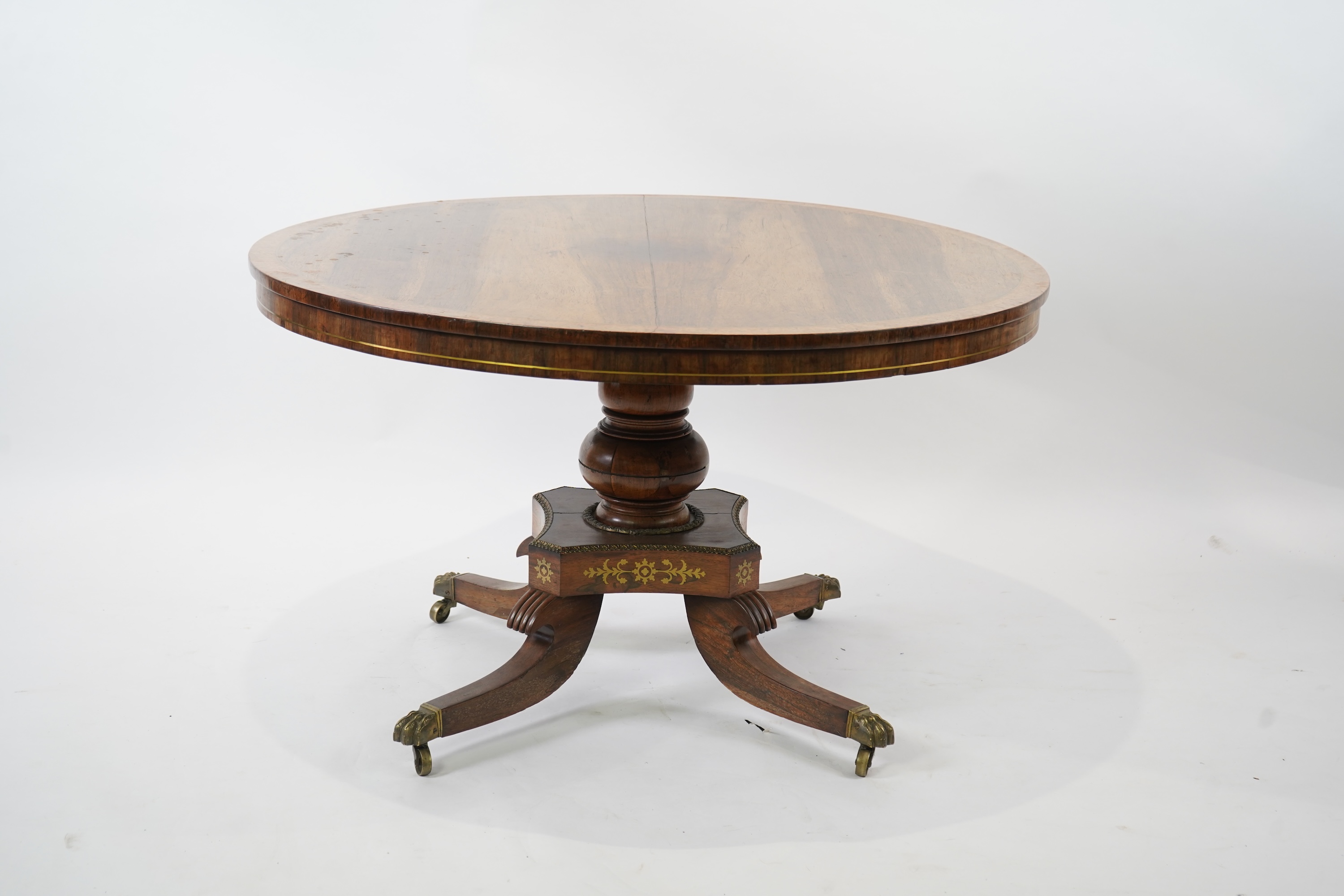 A Regency cut brass inlaid and gilt metal mounted circular rosewood breakfast table, the bird's-eye maple banded tilt top on baluster column, concave platform and quadruple downswept base with cast claw caps and castors,